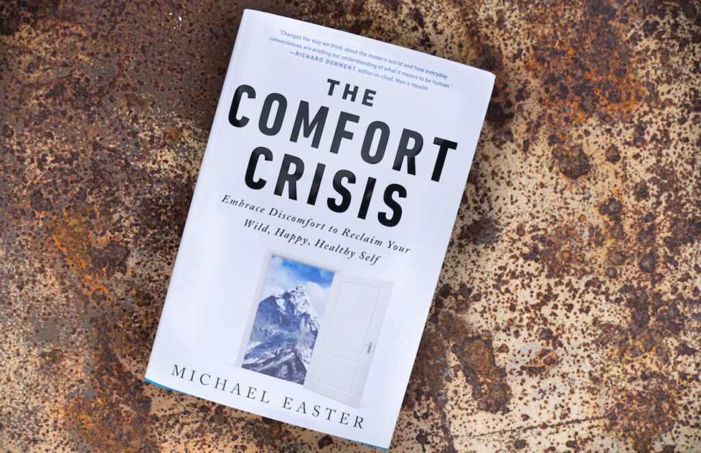 Photo of the front cover of "The Comfort Crisis" book by Michael Easter. On a rusty metal floor. 