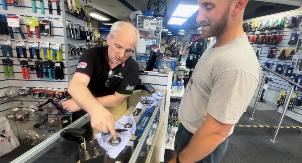 Bike Shop worker behind a counter in his shop, pointing to what parts not to lose, while Louis stands watching. 