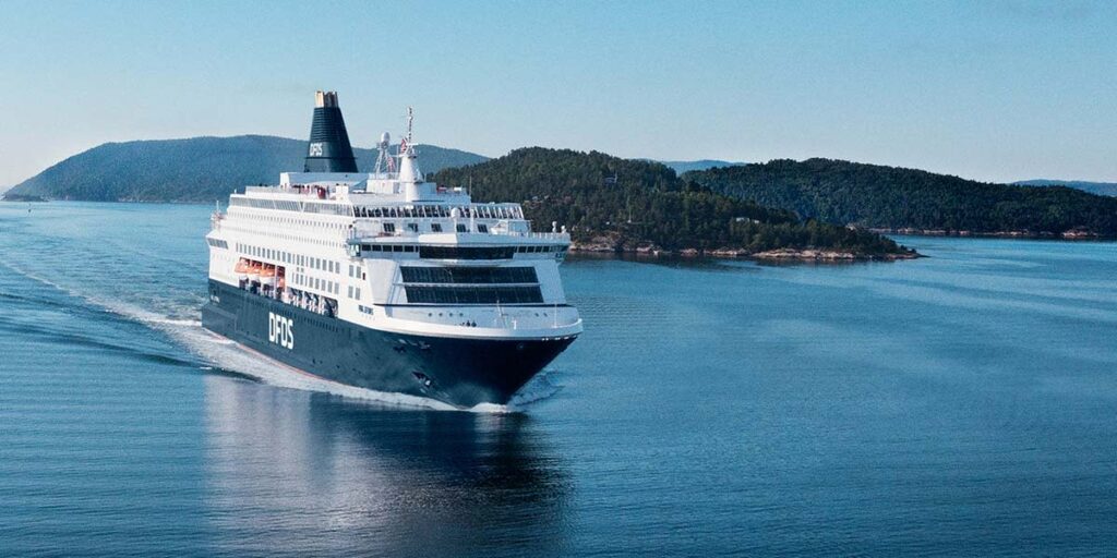 Photo of a DFDS ferry sailing through very calm water, in front of an island with many trees. 